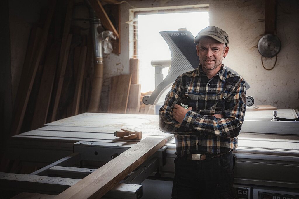 Ein Mann in einem karierten Hemd und einer Mütze steht lächelnd mit verschränkten Armen in einer Holzwerkstatt neben einer Tischsäge und einem Holzbrett. Das Sonnenlicht fällt durch ein Fenster hinter ihm und erhellt den Arbeitsbereich.