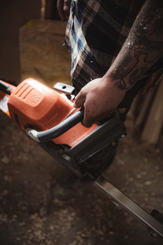 Eine Person mit einem tätowierten Arm hält eine orangefarbene Kettensäge in einem Raum, in dem Sägemehl auf dem Boden liegt.