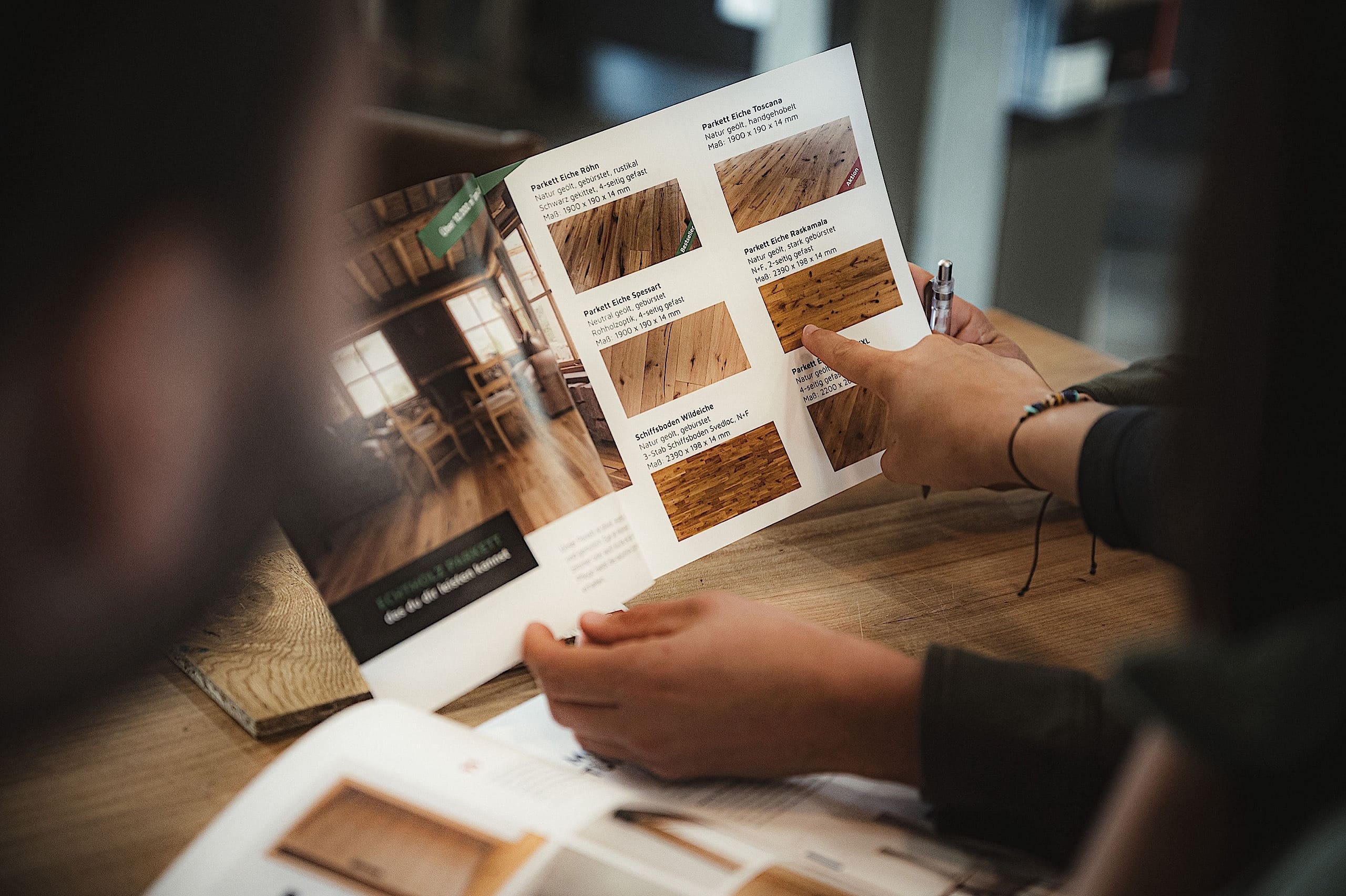 Zwei Personen sitzen an einem Tisch und sehen sich eine Broschüre mit Mustern und Beschreibungen von Holzfußböden an, wobei eine Person mit einem Stift auf die verschiedenen Möglichkeiten hinweist.