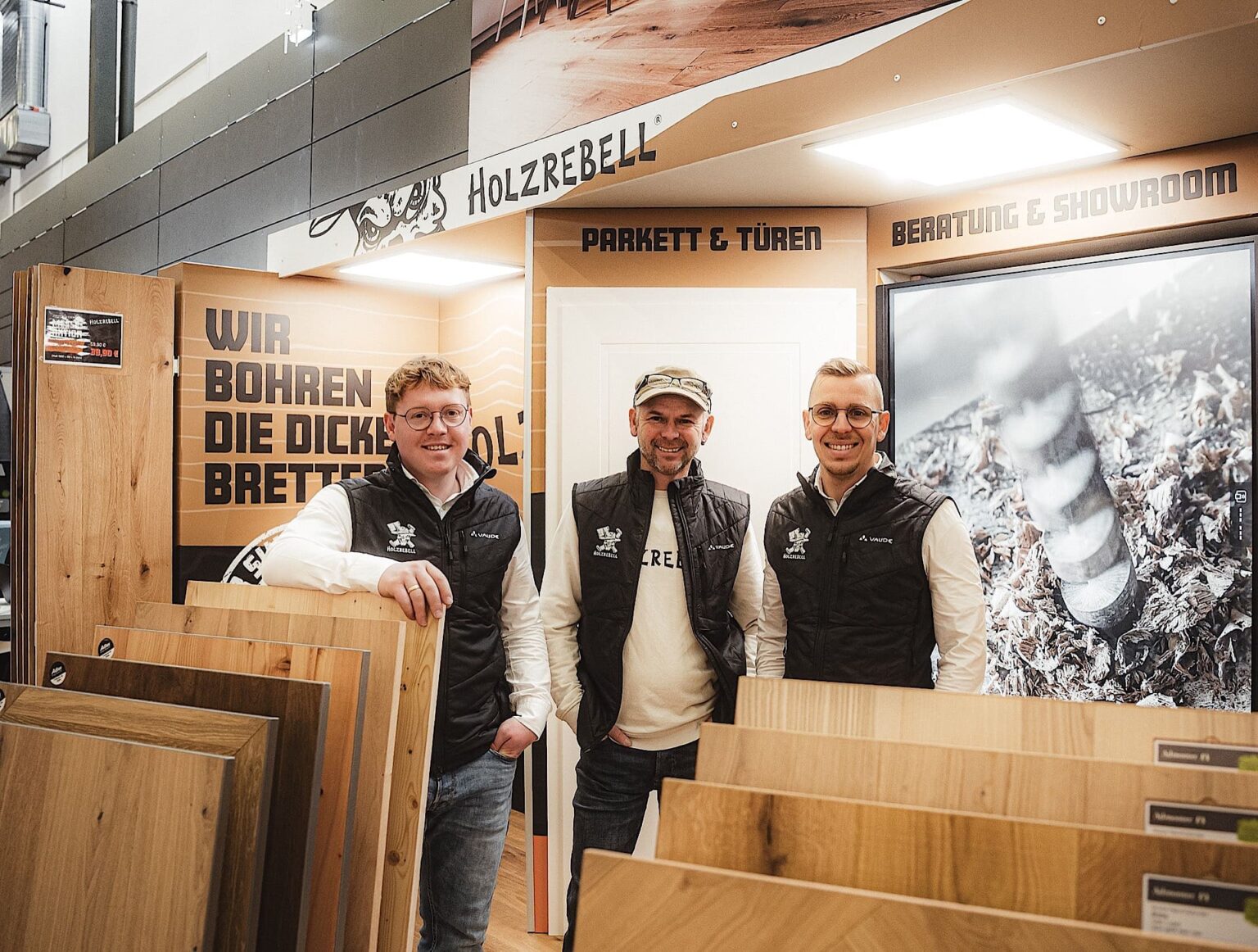 Drei Männer stehen in einem Ausstellungsraum mit Mustern von Holzfußböden, und an den Wänden hinter ihnen sind Schilder in deutscher Sprache über Parkett, Türen und Beratungsdienste zu sehen.