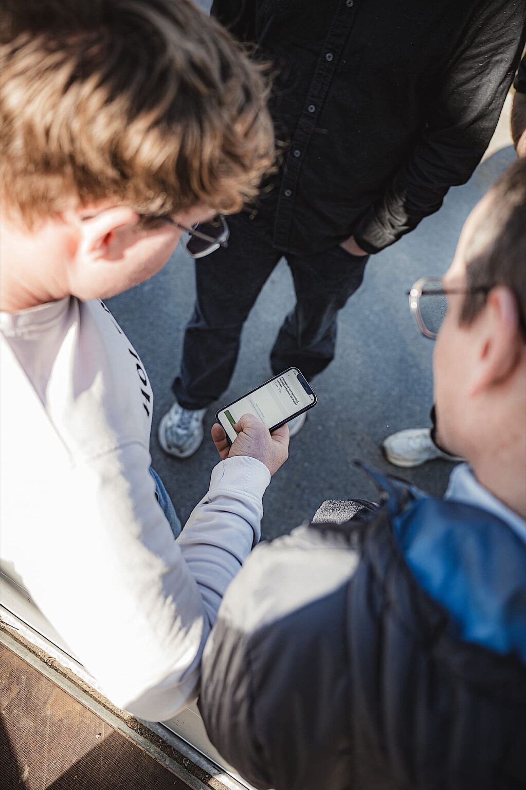 Zwei Personen stehen im Freien, während die eine ein Smartphone in der Hand hält und der anderen eine Nachricht oder einen Text auf dem Bildschirm zeigt, wobei eine dritte Person teilweise im Hintergrund zu sehen ist.