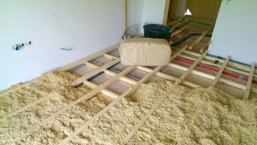 Holzbalken, die mit Schüttmaterial in einem zu renovierenden Raum gedämmt werden; ein Ballen Dämmmaterial und Bauwerkzeuge sind zu sehen.