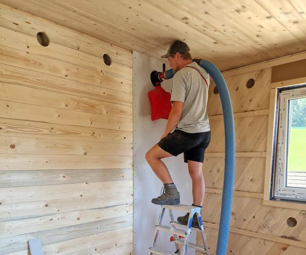 Eine Person steht auf einer Trittleiter und benutzt einen großen Schlauch, um die Wand eines im Bau befindlichen Holzraums zu isolieren.