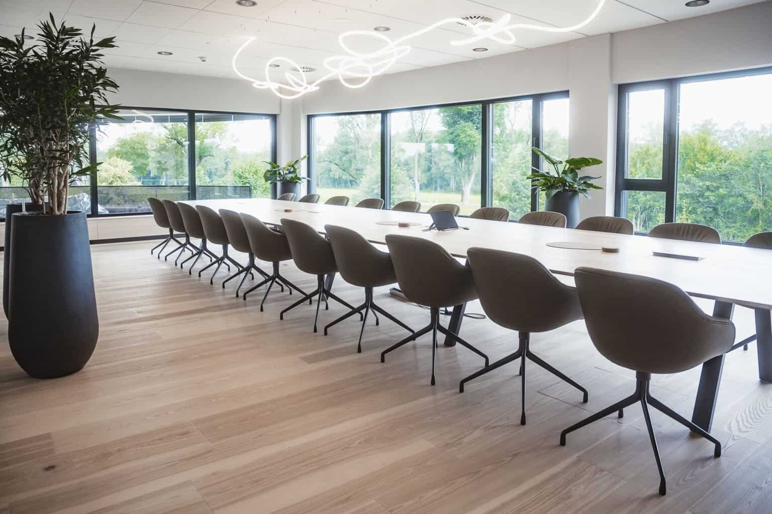 Ein langer Konferenztisch, umgeben von grauen Drehstühlen, in einem hellen Büro mit raumhohen Fenstern.