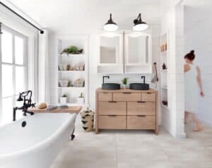 Ein helles Badezimmer mit einer weißen freistehenden Badewanne, einem doppelten Waschtisch aus Holz mit schwarzen Aufsatzwaschbecken und Einbauregalen.