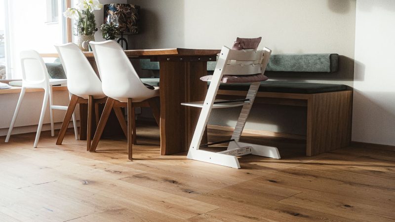 Ein moderner Essbereich mit einem Holztisch, weißen und hölzernen Stühlen, einer Bank mit grünen Kissen und Sonnenlicht, das durch ein Fenster einfällt.