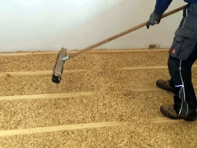 Person, die mit einem Rechen in einem Raum loses Dämmmaterial auf einem Holzbodenrahmen ausbreitet.