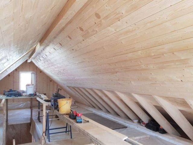 Unausgebauter Dachboden mit Holzwänden und -decke, sichtbaren Dachsparren, Bauwerkzeugen und verstreutem Material.