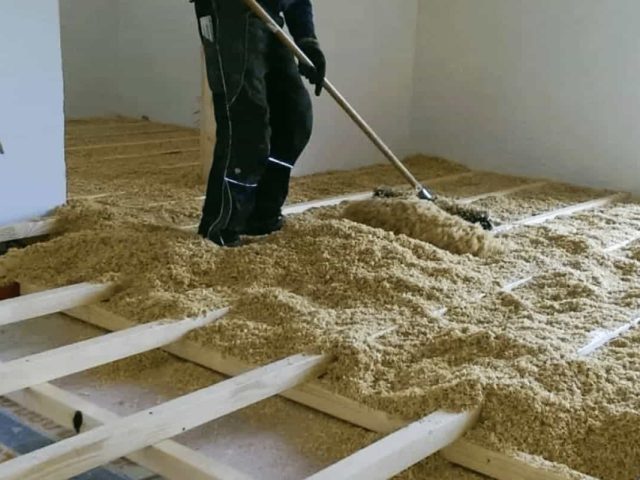 Eine Person verteilt loses Dämmmaterial zwischen Holzbalken in einem Raum, der gerade gebaut oder renoviert wird.