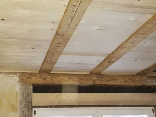 Freigelegte Holzbalken und helle Holzpaneele an einer Decke über einem Fenster in einem unfertigen oder renovierten Raum.