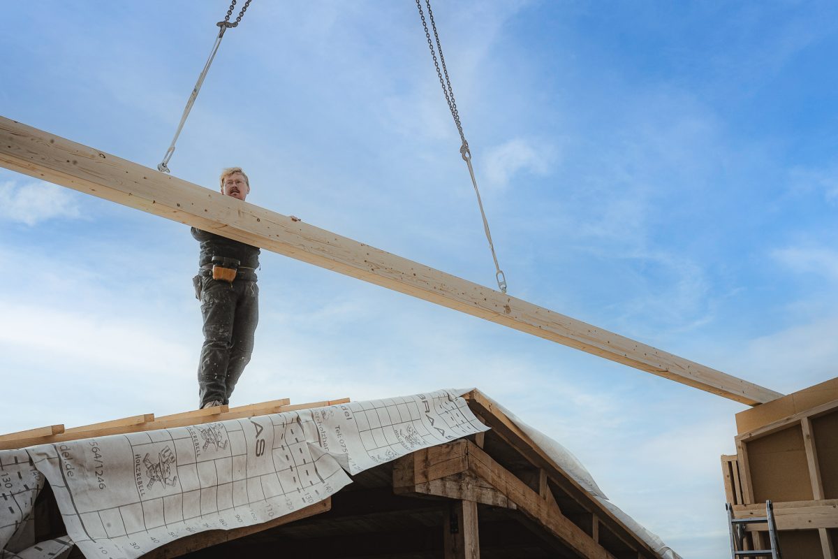 Ein Bauarbeiter steht auf einem Dach, während an einem klaren Tag ein großer Holzbalken von einem Kran angehoben wird. Auf dem Bauwerk darunter sind Baupläne zu sehen.