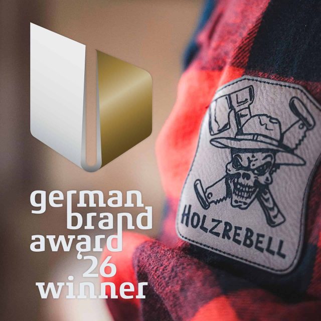 Das Logo des Gewinners des German Brand Award 26 neben einem Holzrebell-Marken-Aufnäher, auf dem ein Totenkopf mit Hut und gekreuzten Äxten abgebildet ist.