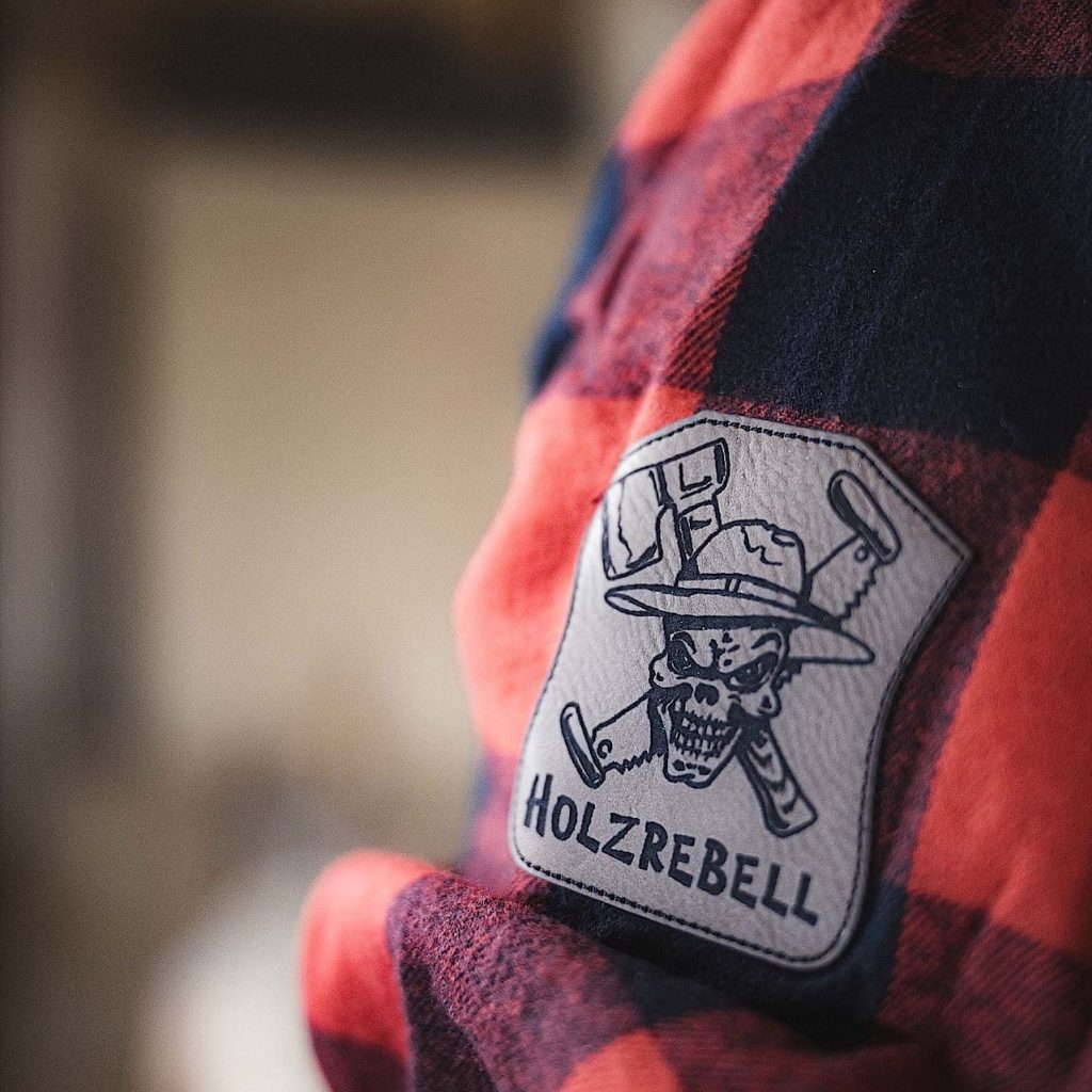 Nahaufnahme eines rot-schwarz karierten Hemdsärmels mit einem Aufnäher mit einem Totenkopf, gekreuzten Werkzeugen und dem Wort HOLZREBELL.