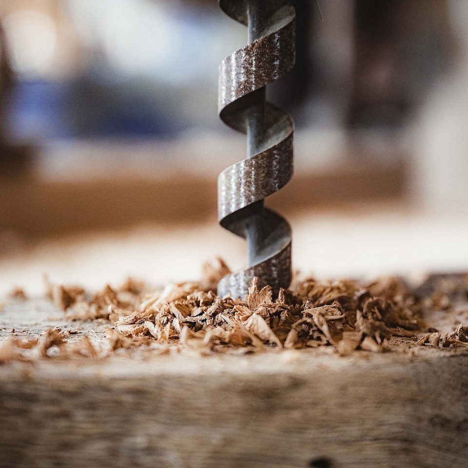 Eine Nahaufnahme eines Bohrers, der sich in ein Stück Holz bohrt, mit Holzspänen, die um das Loch herum verstreut sind. Der Hintergrund ist leicht unscharf und lenkt den Blick auf den Bohrvorgang.