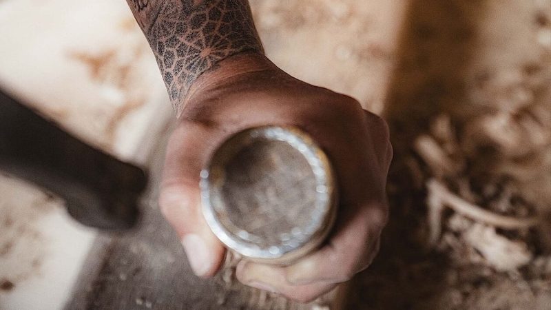 Eine tätowierte Hand ergreift einen Meißel und schnitzt in eine hölzerne Oberfläche, über die Holzspäne verstreut sind.