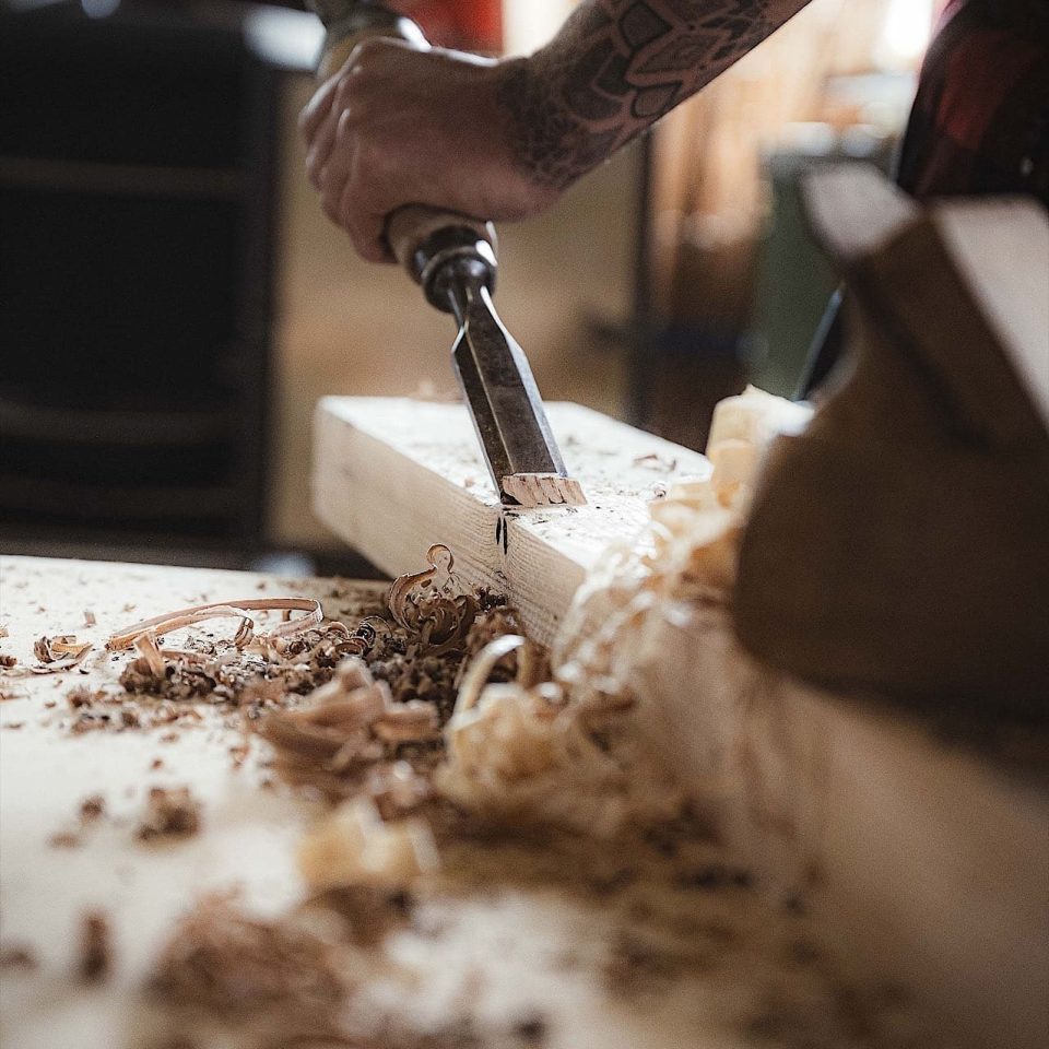 Eine Person schnitzt mit einem Meißel und einem Hammer Holz, während auf einer Werkbank Holzspäne verstreut sind.