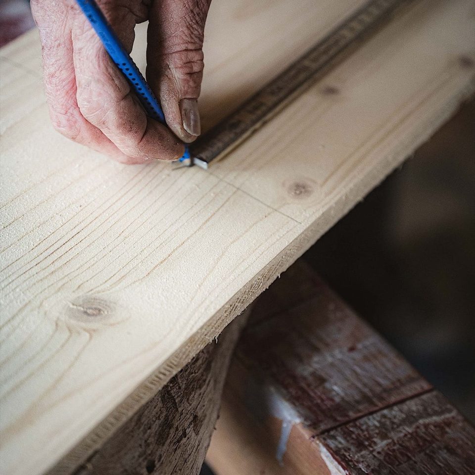 Eine Person benutzt einen Bleistift und ein Lineal, um Maße auf einem Stück hellen Holzes zu markieren.