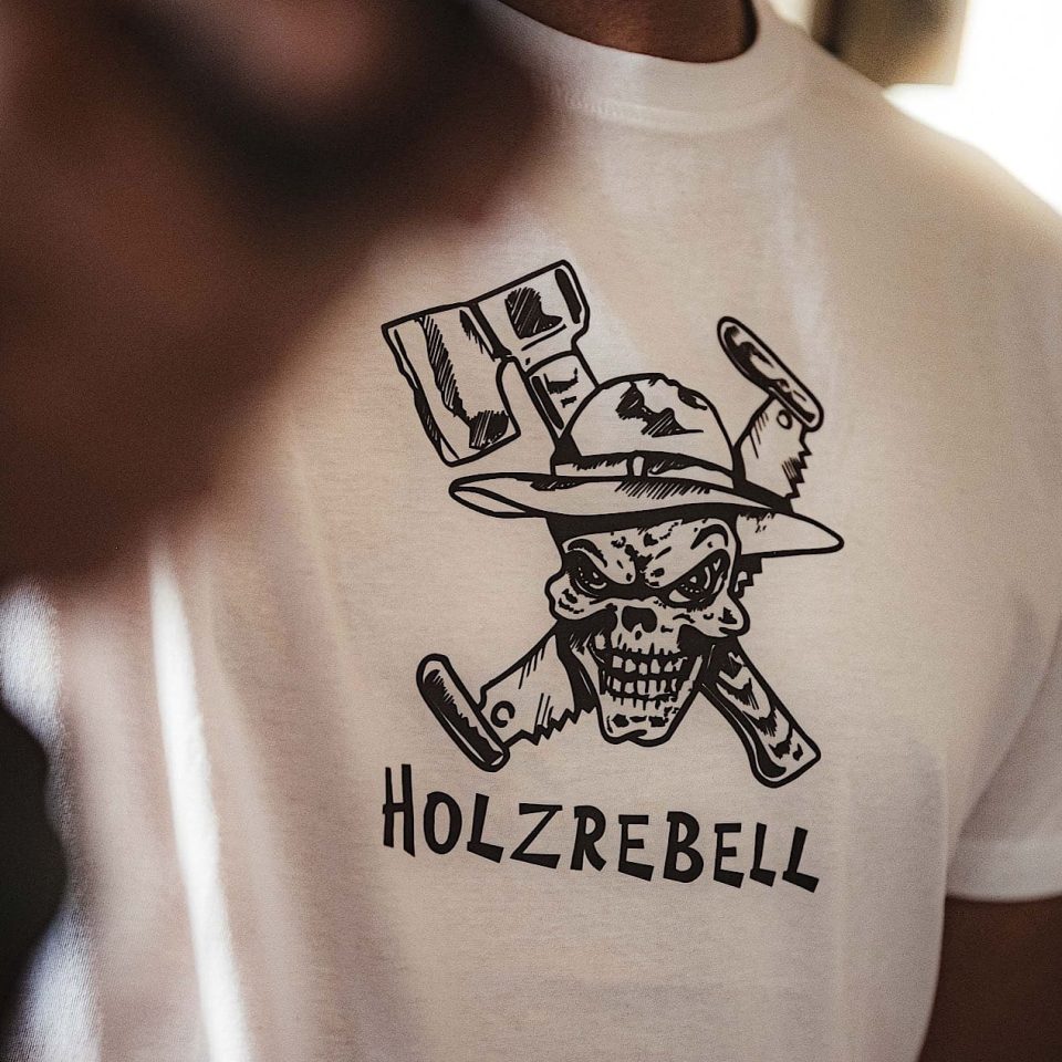 Eine Person, die ein weißes T-Shirt mit der Abbildung eines Totenkopfes mit Hut und gekreuzten Äxten und dem Wort HOLZREBELL darunter trägt.