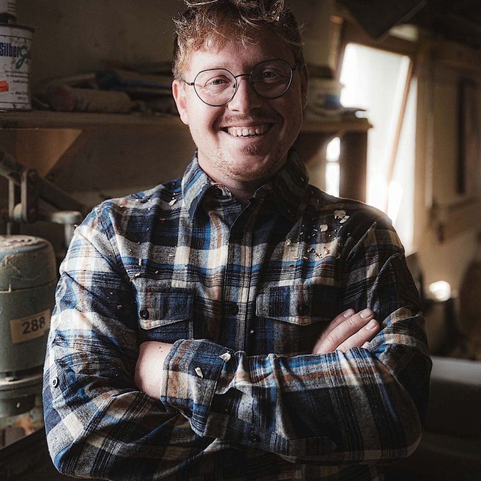 Eine Person mit Brille und kariertem Hemd steht mit verschränkten Armen in einem Haus und lächelt. Im Hintergrund sind Regale und Werkstattausrüstung zu sehen.