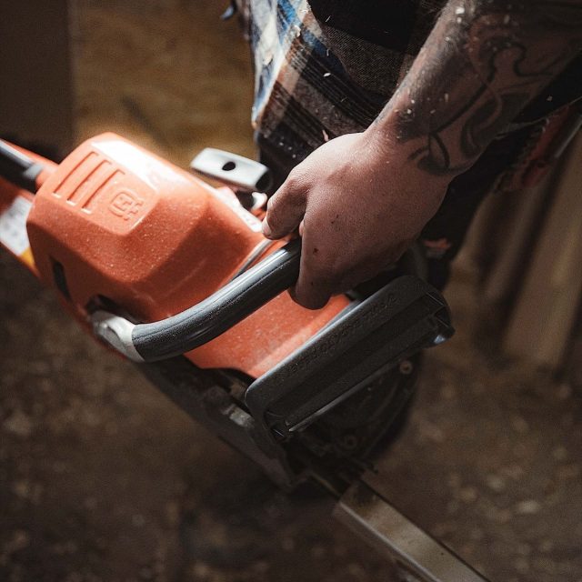 Eine Person mit einem tätowierten Arm hält eine orangefarbene Kettensäge in einem Raum, in dem Sägemehl auf dem Boden liegt.