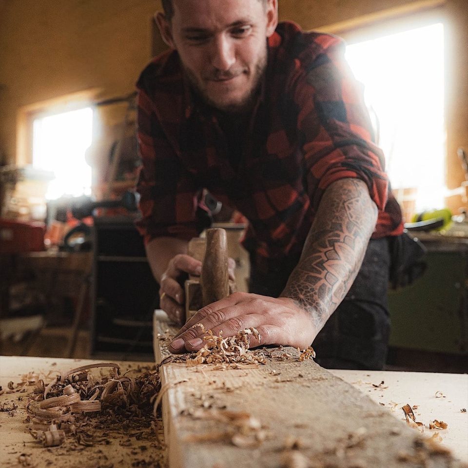 Ein Mann in einem rotkarierten Hemd hobelt in einer Werkstatt mit einer Handhobelmaschine ein Holzbrett ab. Holzspäne bedecken die Werkbank, und Sonnenlicht fällt durch das Fenster hinter ihm.