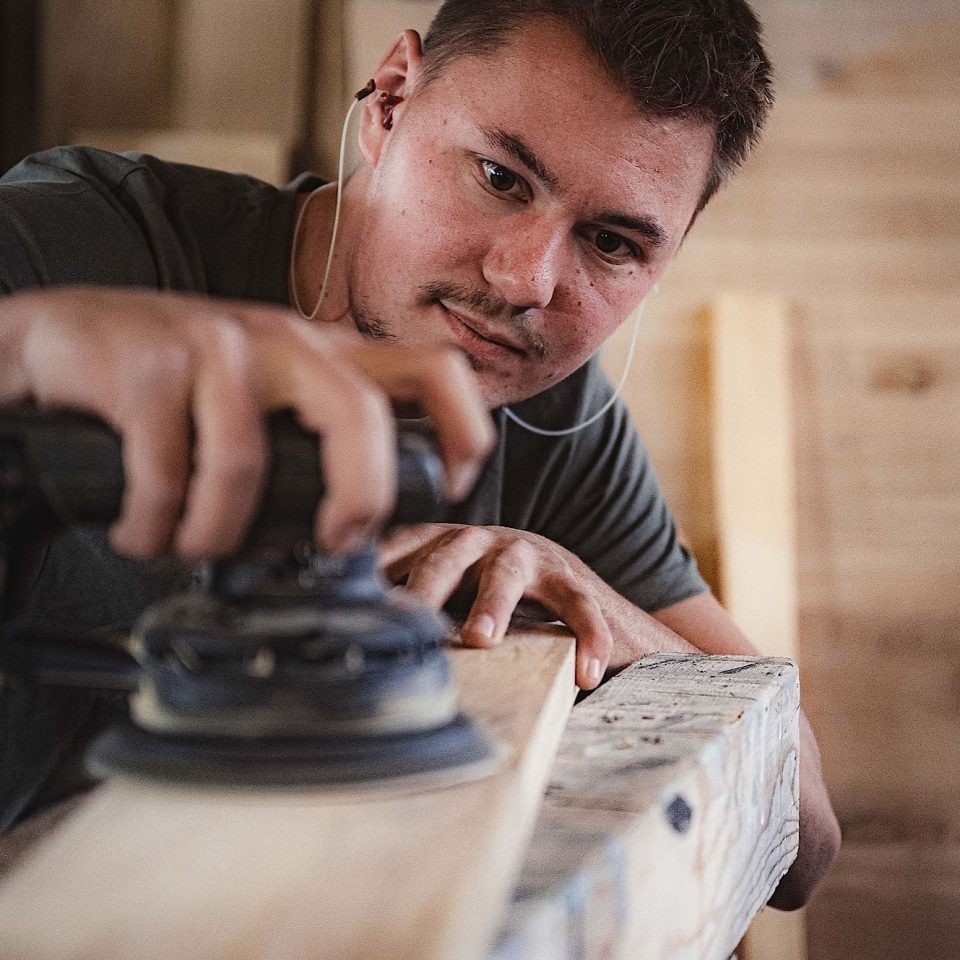 Eine Person benutzt eine elektrische Schleifmaschine, um ein Holzbrett in einer Werkstatt zu glätten.
