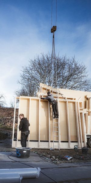 Zwei Arbeiter montieren im Freien große Holzwandplatten; einer steht auf einer Leiter und justiert eine von einem Kran aufgehängte Platte, während der andere in der Nähe steht und Werkzeuge hält.