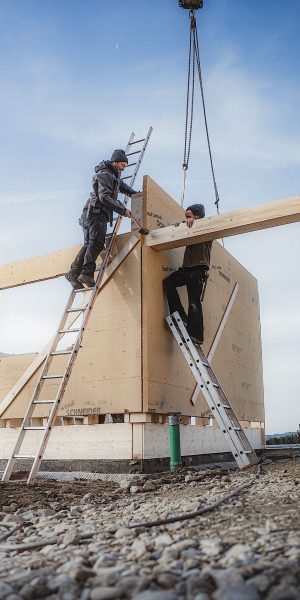 Zwei Bauarbeiter auf Leitern führen einen großen Holzbalken, der von einem Kran auf einer Baustelle mit einer Holzrahmenkonstruktion angehoben wird.