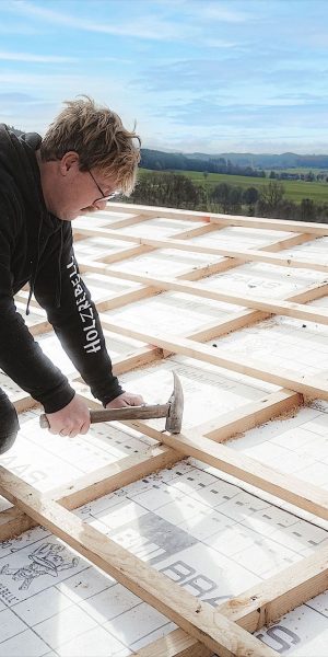 Eine Person kniet auf einem Dachstuhl und befestigt mit einem Hammer Holzlatten über der Isolierung. Im Hintergrund ist eine ländliche Landschaft zu sehen.