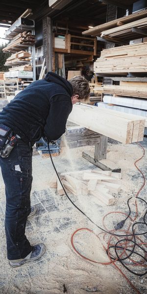 Eine Person in Arbeitskleidung schneidet mit einem Elektrowerkzeug einen Holzbalken im Freien, umgeben von Holz und Sägemehl.