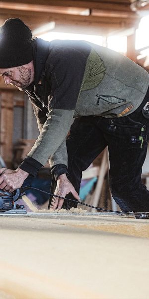 Eine Person schneidet mit einer Kreissäge Holz in einer Werkstatt mit Holzstapeln im Hintergrund.