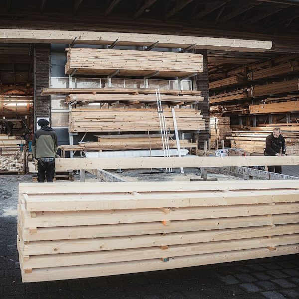 Arbeiter hantieren mit gestapelten Holzbalken vor einem Holzlager, während im Vordergrund ein Gabelstapler eine Palette mit Holz bewegt.