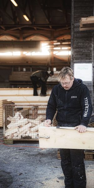 Eine Person in Arbeitskleidung misst und markiert einen Holzbalken in einer Holzwerkstatt; eine andere Person arbeitet im Hintergrund in der Nähe von gestapeltem Holz.