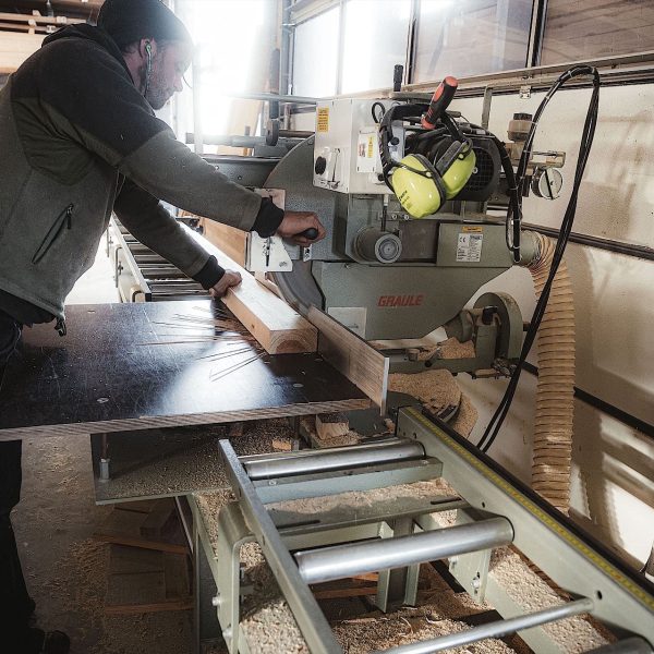 Eine Person bedient in einer Werkstatt eine Holzbearbeitungsmaschine und schneidet ein Stück Holz zu; in der Nähe sind Schutzkleidung und Werkzeuge zu sehen.