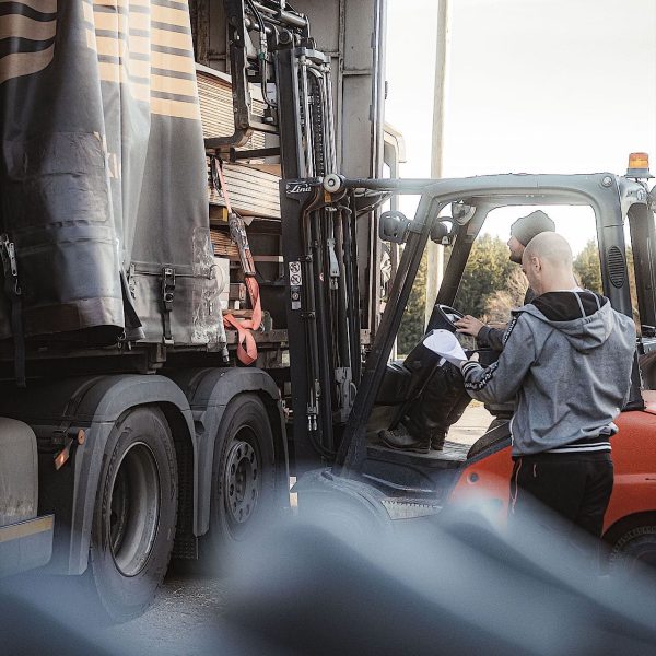 Ein Mann steht neben einem Gabelstapler und einem Lastwagen, hält ein Klemmbrett in der Hand und prüft Dokumente, während der Gabelstapler Materialien auf den Lastwagen lädt oder von ihm ablädt.
