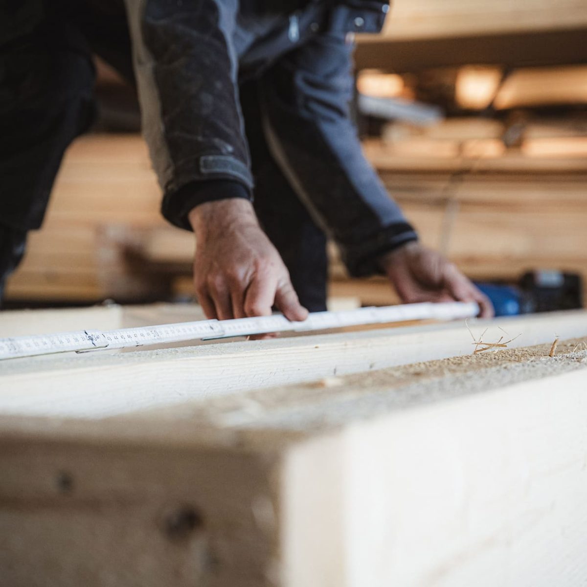 Eine Person misst in einer Werkstatt ein Holzbrett mit einem Maßband aus, wobei sie sich auf ihre Hände und das Holz konzentriert.