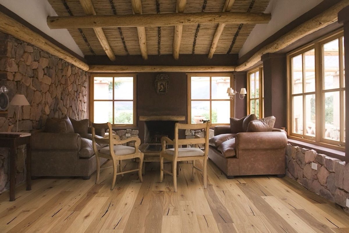 Rustikales Wohnzimmer mit Steinwänden, hölzernen Deckenbalken, großen Fenstern, Ledersofas und Holzstühlen, die um einen zentralen Tisch auf Hartholzböden angeordnet sind.