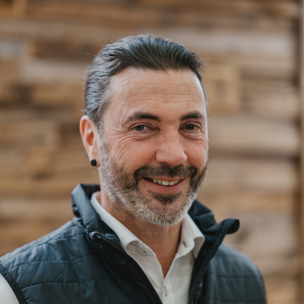 Mann mittleren Alters mit kurzen dunklen Haaren und Bart, der ein weißes Hemd und eine dunkle Weste trägt, lächelt vor einem hölzernen Hintergrund.