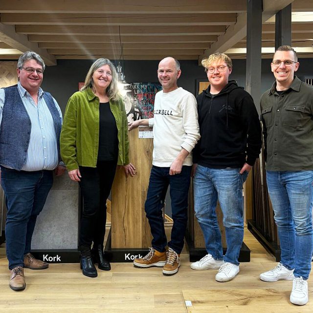 Fünf Personen stehen drinnen auf einem Holzboden und posieren für ein Gruppenfoto vor Schautafeln mit der Aufschrift Kork.
