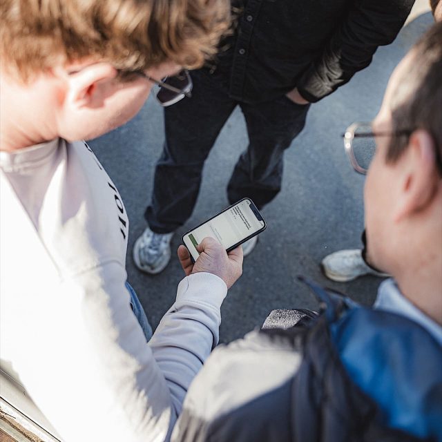 Zwei Personen stehen im Freien, während die eine ein Smartphone in der Hand hält und der anderen eine Nachricht oder einen Text auf dem Bildschirm zeigt, wobei eine dritte Person teilweise im Hintergrund zu sehen ist.