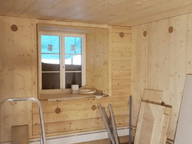 Ein Raum im Bau mit hölzernen Wänden und Decken, einem Fenster in der Mitte, Baumaterialien und Werkzeugen, die auf dem Boden verstreut liegen.